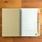 Open spiral notebook with lined pages and orange pen on a wooden table.
