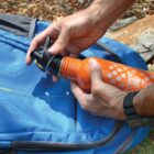 Person placing an orange water bottle into a blue backpack outdoors.
