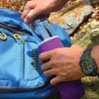 Hands placing a purple-cased water bottle into a blue backpack outdoors.