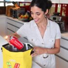 Smiling woman placing a red can into a yellow cooler bag in a modern kitchen.