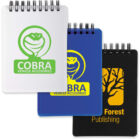 Three spiral notepads: white and blue with Cobra Vehicle Accessories logo, black with Forest Publishing logo.