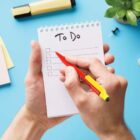 Handwriting a to-do list on a notepad with a red pen, surrounded by office supplies on a blue surface.