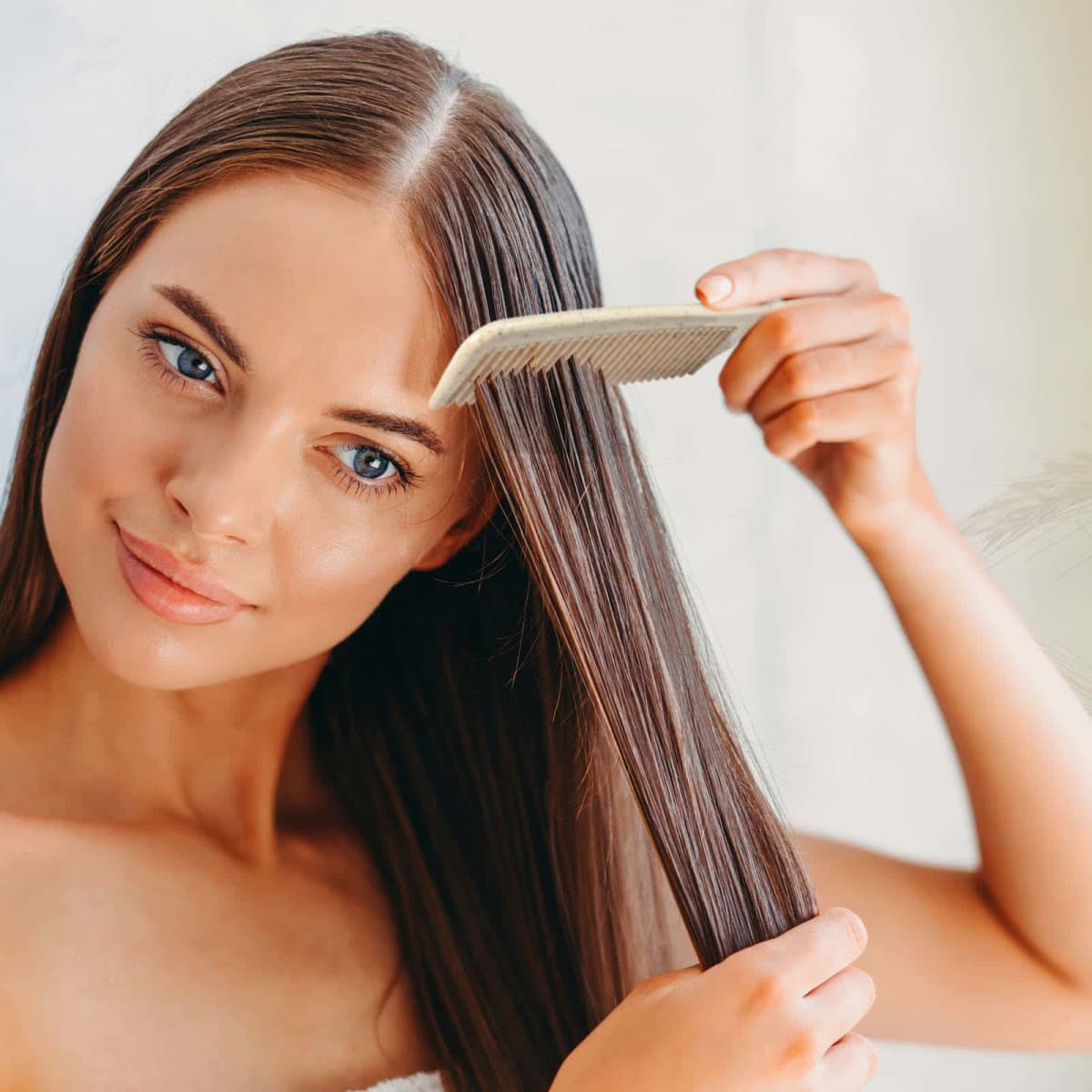 Woman with long brown hair using a comb, smiling and looking to the side, against a light background.