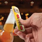 A hand opening a bottle of beer with a bottle opener, against a blurred background with warm bokeh lights.