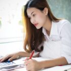 Person in white shirt working on a laptop, writing with a red pen, looking focused.