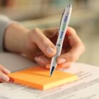 A hand writes on an orange sticky note with a Viva Eco pen.