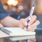 Hand holding a pen poised over an open notebook on a reflective surface.
