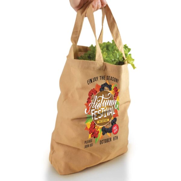 A hand holds a beige tote bag with "Autumn Festival" text, autumn leaves, and green veggies.