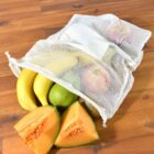 A reusable white mesh bag on a wooden surface contains bananas, a green pear, and apples, with sliced cantaloupe in front of it.