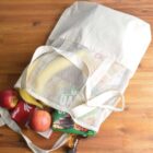 A reusable mesh shopping bag on a wooden surface, filled with grocery items including apples, bananas, a box of Kellogg’s cereal, and other packaged goods. The bag is open, revealing its contents.