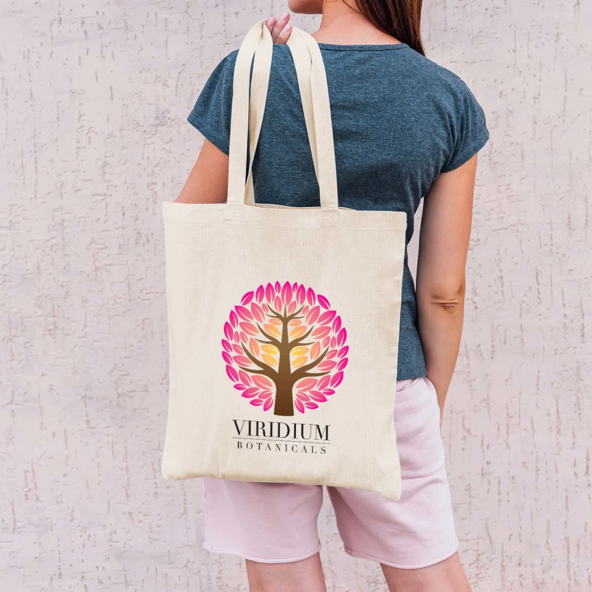 A person in a blue shirt and pink shorts holds a beige tote bag over their shoulder. The bag features a colorful tree design with pink, orange, and yellow leaves and text that says Viridium Botanicals.