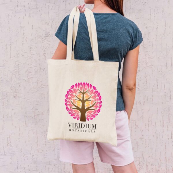 A person wearing a blue shirt and pink shorts is holding a beige tote bag with a colorful tree logo and the text Viridium Botanicals against a textured white wall.