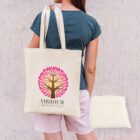 A person with long hair, wearing a blue shirt and pink shorts, carries a tote bag with a colorful tree logo and the text Viridium Botanicals. A folded tote bag is placed beside them. The background is a textured wall.