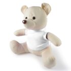 A plush beige teddy bear is sitting upright, wearing a plain white T-shirt. The bear has a round, soft body, small black eyes, and a brown nose, against a plain white background.