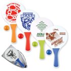 A set of four colorful beach paddles with different designs, accompanied by orange balls. Designs include NETWORK RECRUITS, HUNGRY BEARS 2016, AQUA RESORT, and WINGSUIT FLYING QUEENSTOWN NEW ZEALAND. A black mesh bag is also included.