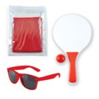 A set of beach items: red sunglasses with dark lenses, a red paddle and ball set, and a red mesh bag in a plastic pouch. The paddle has a white surface with a red handle.