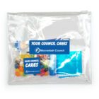 A clear zippered bag containing candy, a blue foil packet, a small toy, and a labeled hand sanitizer bottle. Two blue stickers on the bag read Your Council Cares and Mooranbah Council.
