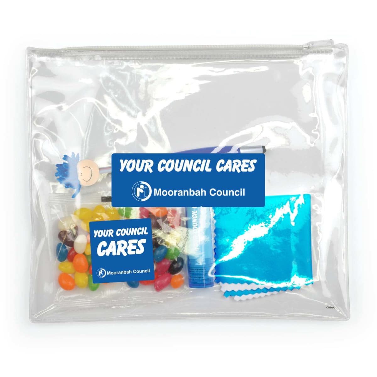 A clear zippered bag containing candy, a blue foil packet, a small toy, and a labeled hand sanitizer bottle. Two blue stickers on the bag read Your Council Cares and Mooranbah Council.