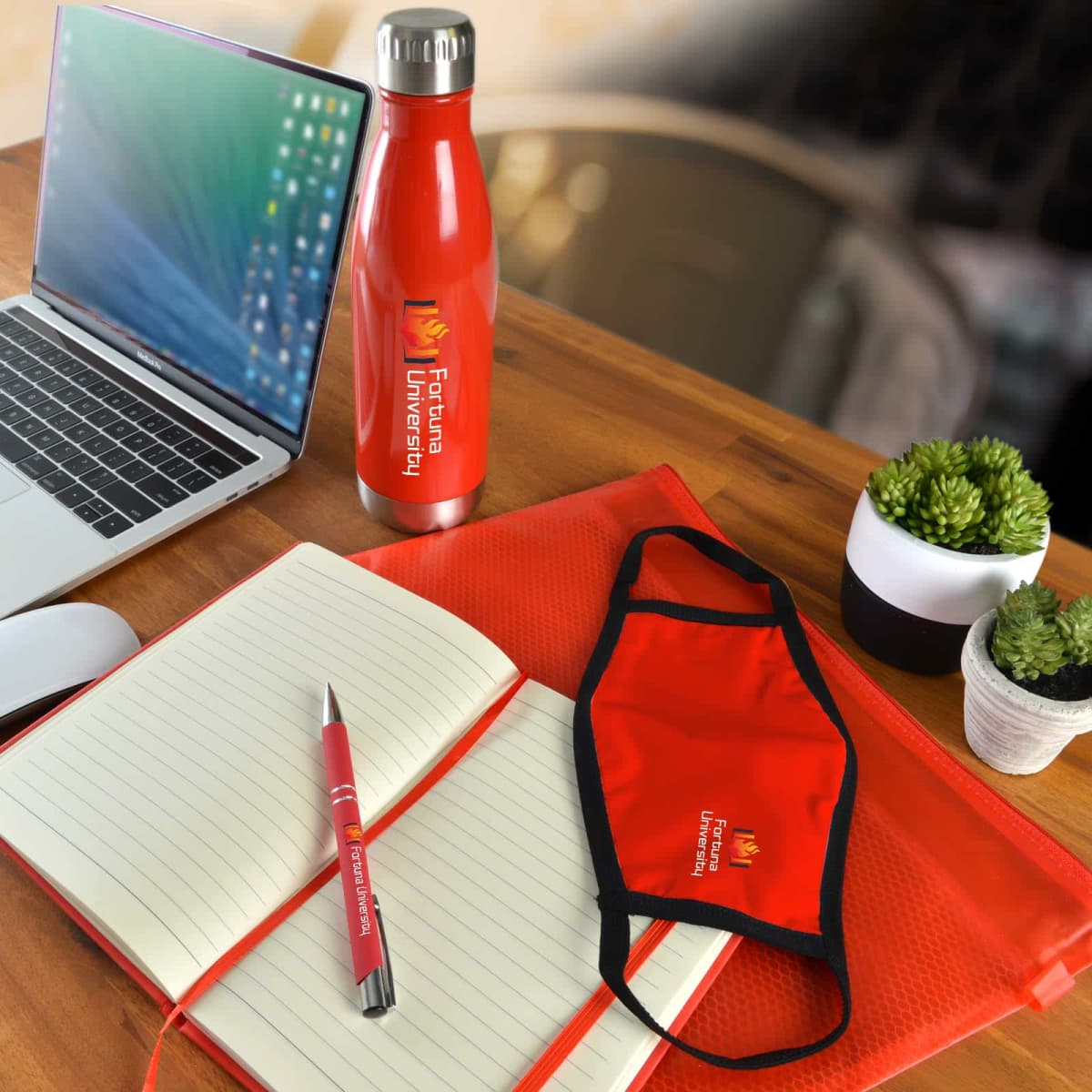 A workspace with a laptop, a red pen, a red water bottle, a red folder, a red face mask, and a notebook, all branded with Forward University. Two small potted plants are nearby on the wooden desk.