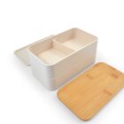 A beige, two-tiered lunchbox with a wooden lid is shown. The top layer features a divider, and both layers are stackable. The lid is placed to the side.