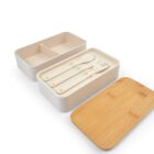 A set of eco-friendly lunch containers with a bamboo lid. One container is open, showing a fork, spoon, and knife inside. Another container is empty with a divider. The bamboo lid is placed leaning against the containers.