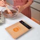 A person in a pink shirt mixes chocolate batter in a glass bowl with a wooden spoon on a white countertop. An electronic kitchen scale with a bamboo surface is in the foreground.