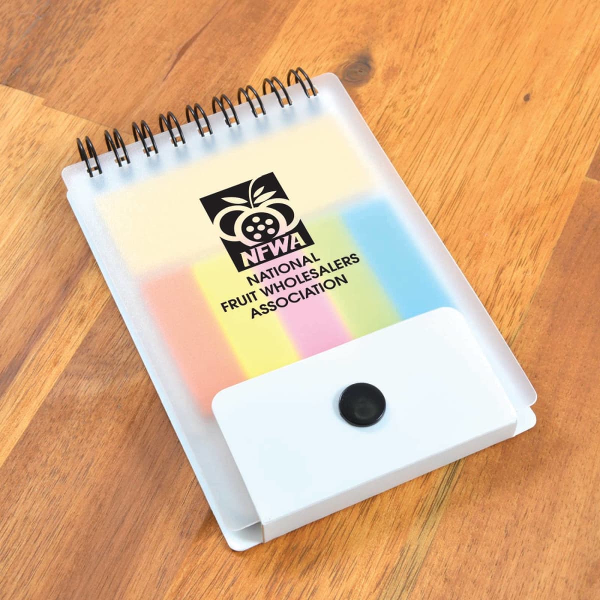 A small spiral notepad with a translucent cover lies on a wooden surface. The cover features the logo of the National Fruit Wholesalers Association, depicted as an apple with the acronym NFWA above the full name, and has a snap closure at the bottom.