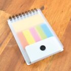 A small spiral notepad with a transparent cover lies on a wooden surface. Inside, there are sticky notes in yellow, pink, green, and blue. A black button clasp is visible on the cover.