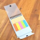 A small spiral notepad with a transparent cover is open on a wooden surface. Inside, there are sticky notes in various colors: yellow, orange, pink, green, and blue. The notepad is positioned at an angle.