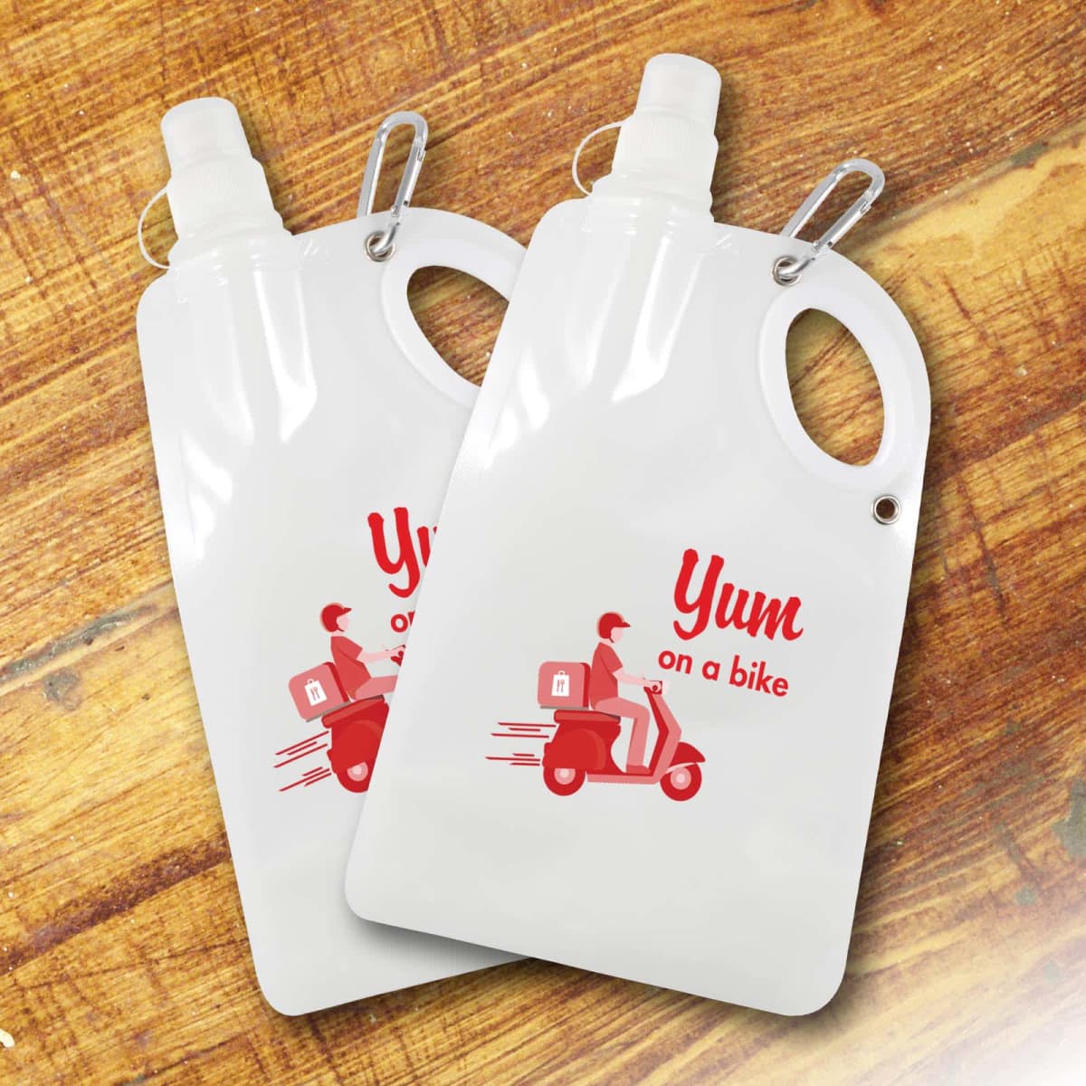 Two collapsible water bottles with carabiners on a wooden surface. Each features a red graphic of a delivery person on a scooter with the text Yum on a bike.