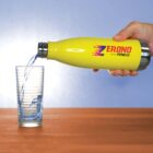 A hand pours water from a yellow stainless steel bottle labeled Zerono Fitness into a clear glass. The glass is on a wooden surface, and the background is a light gradient of blue and white.