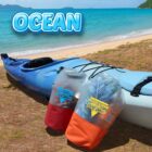 Blue kayak on a sandy beach with calm ocean waves. Two waterproof bags, red and orange, labeled Pipeline Anywhere are attached to the kayak. Blue sky and distant islands are visible in the background, with the word OCEAN in bold.