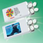 Two white sliding mint containers on a green background. Top container features a colorful flower logo with Life Wealth text. Bottom container reads Immunology Conference with event details. Both containers have white mints partially visible.