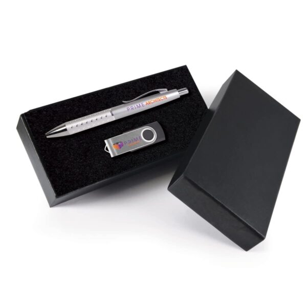 A silver pen and USB flash drive with branding are displayed inside an open black gift box. The pen and USB drive are arranged neatly on a soft black cushion within the box. The box lid is partially visible to the right.