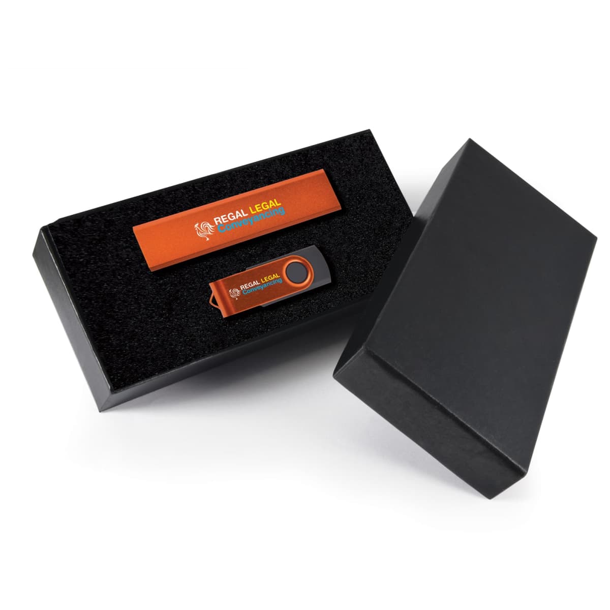 A USB flash drive with an orange swivel design, branded Regal Legal, is displayed inside a black foam-lined gift box. The box also contains an orange pen with the same branding. The box lid is leaning against its side.