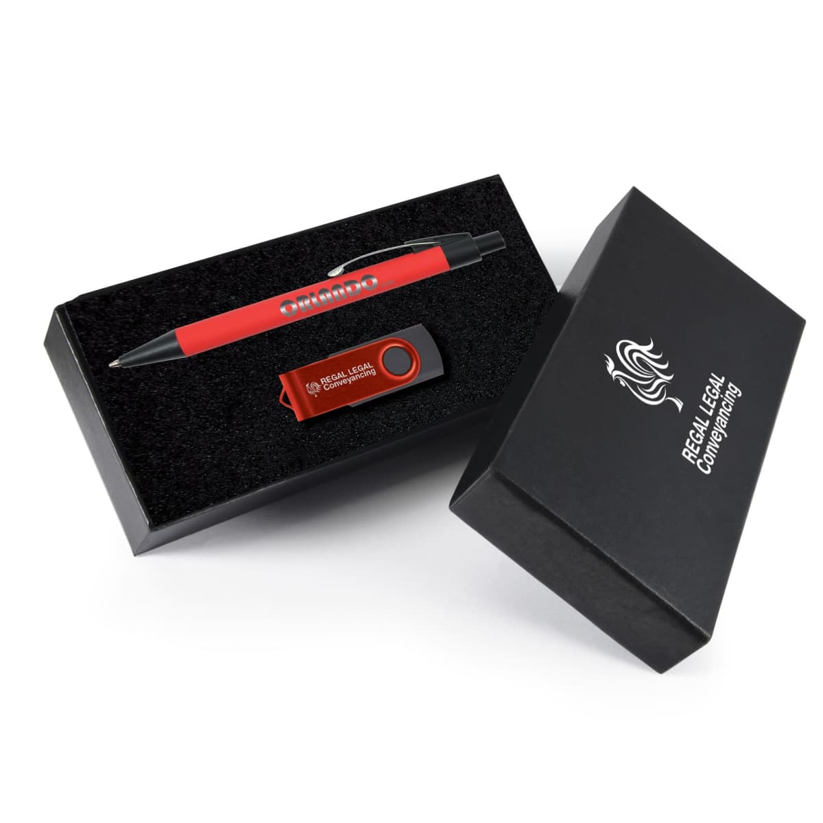 A black gift box with open lid contains a red ballpoint pen and a red USB flash drive. The box has a logo and text on the side. The pen and USB are neatly placed in black foam.