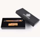 A black gift box with a logo on the lid is displayed open, revealing a bamboo pen and a USB drive inside. Both items have the word ADVANTAGE printed on them, with the USB also featuring a small apple design.