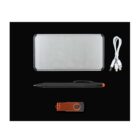 A flat lay of office accessories on a black background, including a silver rectangular power bank, a white three-in-one charging cable, a black pen with an orange tip, and an orange USB drive.
