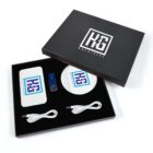 A branded gift set in a black box, containing two wireless chargers with a logo, a blue USB drive, and two white charging cables, all neatly arranged on a black background. The box lid displays the same logo.