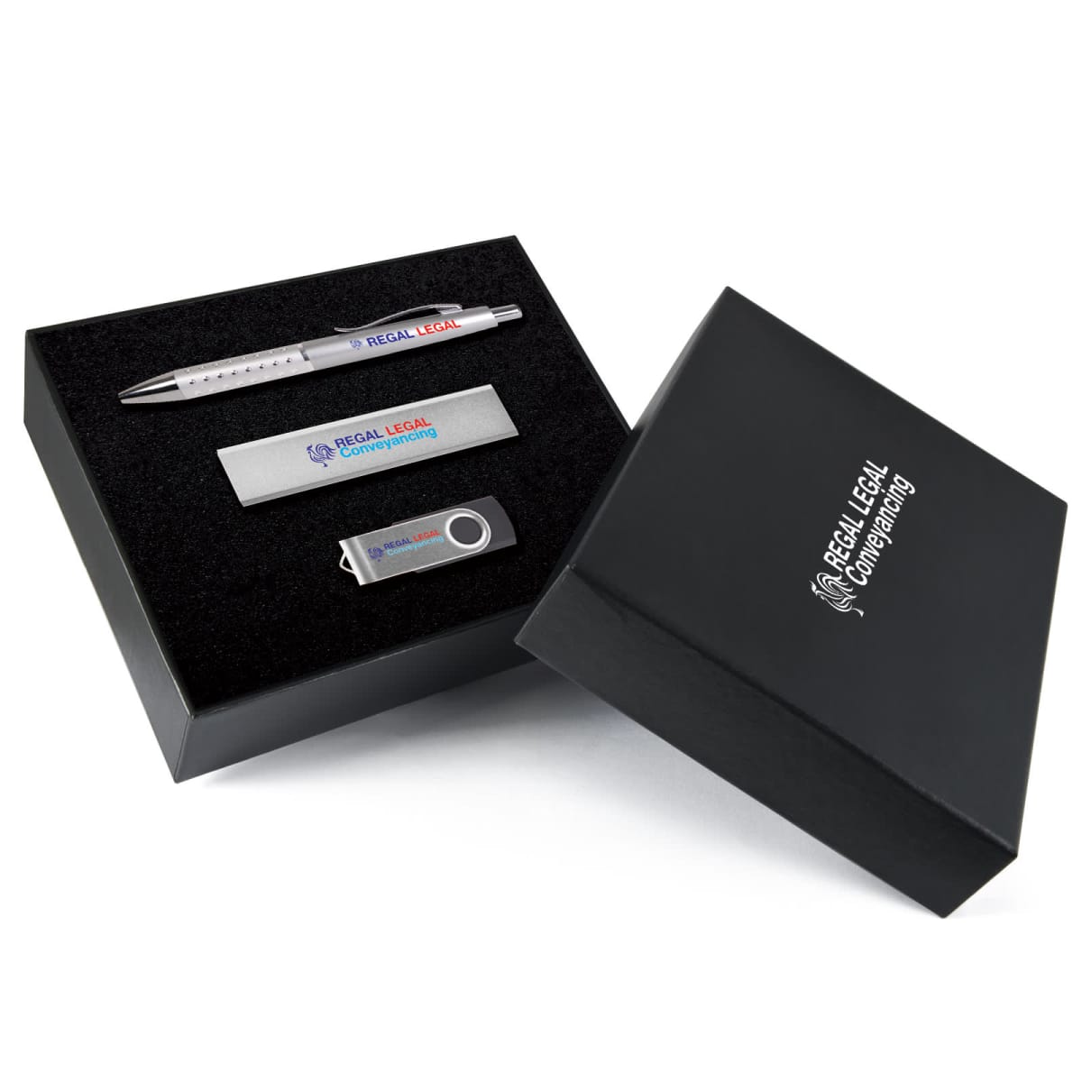 A black gift box with a hinged lid containing a silver pen, a silver USB flash drive, and a rectangular silver metal object, each engraved with REGAL LEGAL. The box is labeled REGAL LEGAL COMPANY NAME in white lettering.