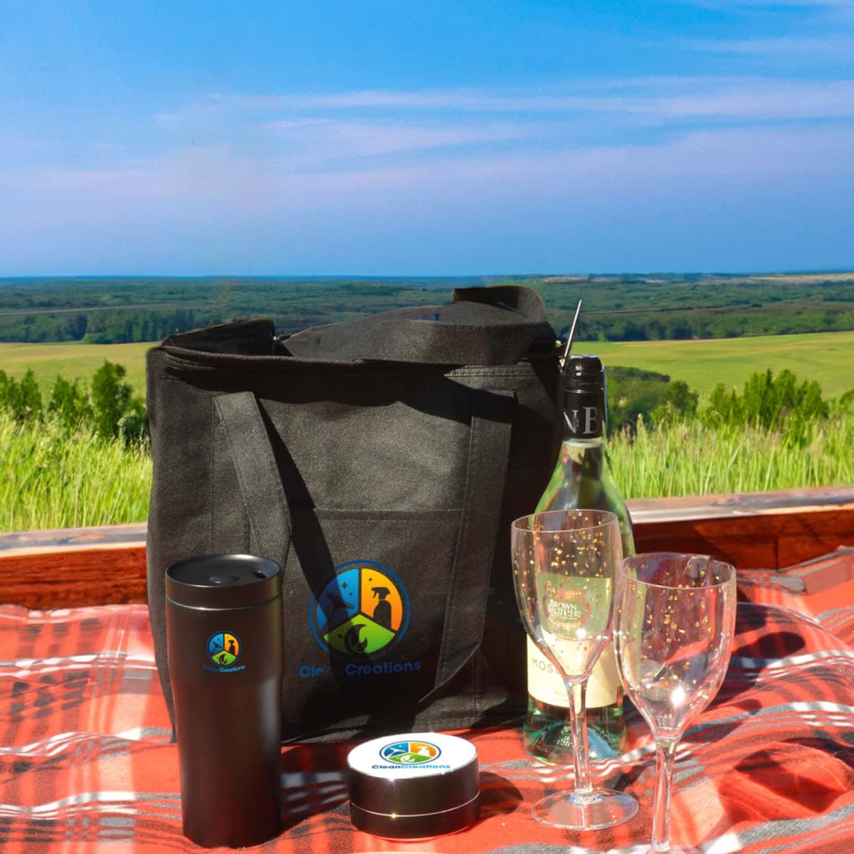 A picnic setup on a plaid blanket features a tote bag, two glasses, a wine bottle, and a thermos, all branded with a colorful logo. The background shows a scenic view of lush green fields and a distant blue sky.