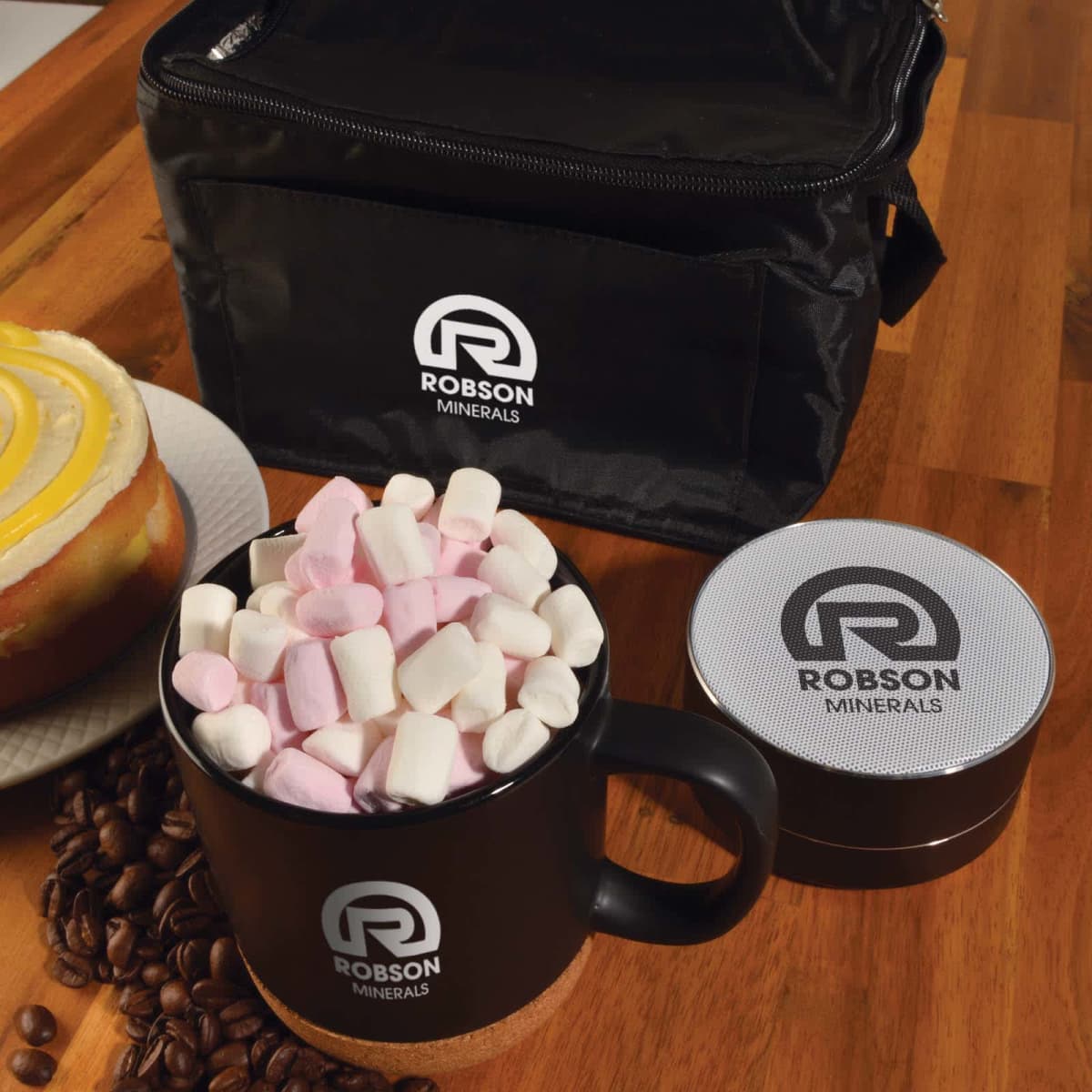 Black mug filled with pink and white marshmallows on a wooden table. A round cake, coffee beans, a round branded container, and a black bag with the Robson Minerals logo are also visible.