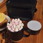 A black mug filled with pink and white marshmallows sits on a wooden table, surrounded by coffee beans. A cinnamon roll on a plate and a portable speaker are nearby, with a black lunch bag in the background.