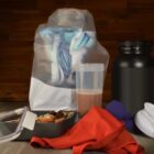 A protein shake in a clear bottle, a black container, a bag with a blurred design, a small meal in an open container, a red towel, purple dumbbells, and a white shaker lid on a wooden surface with a dark wood background.