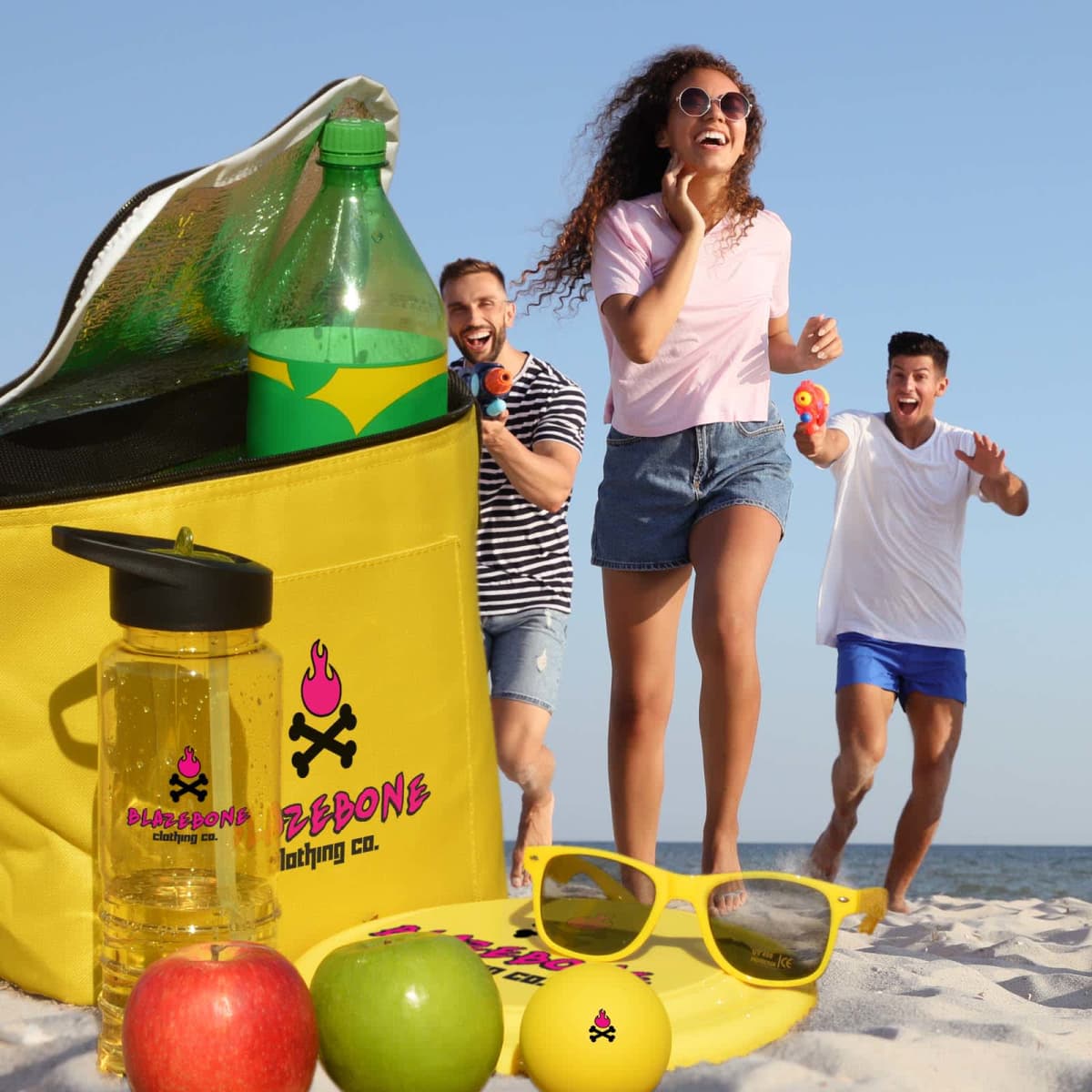 A group of three people enjoy a beach day, smiling and playing with toys. A yellow cooler bag, soda bottle, fruit, plastic cup, and sunglasses are in the foreground. The sand and ocean create a sunny and relaxed backdrop.