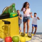 Three people enjoy a sunny beach day, laughing and playing with toys. A yellow cooler with a green bottle, apples, a yellow ball, and sunglasses sits in the sand nearby. The sea is visible in the background.