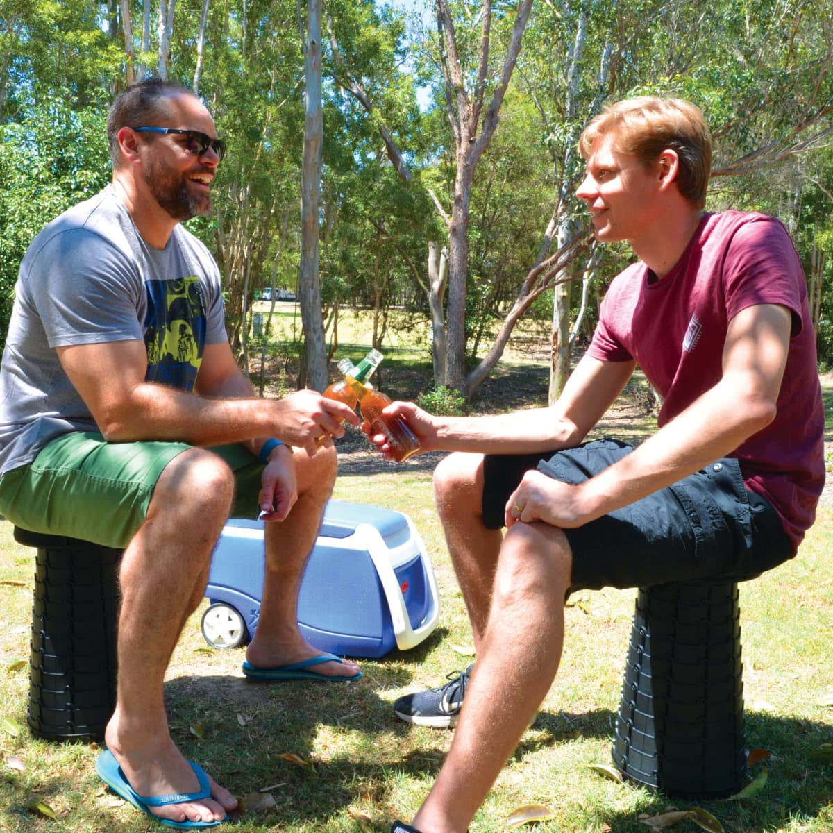 Two men sit outdoors on stackable stools, smiling and clinking beer bottles. A cooler is placed on the grass near them. Theyre surrounded by trees, and the scene conveys a relaxed, casual atmosphere.