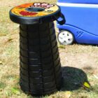 Collapsible plastic stool labeled BBQ Mate on grass next to a blue cooler with wheels. The stool is black with a round top and appears portable.