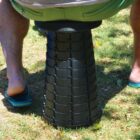 A person wearing green shorts and blue flip-flops sits on a black, collapsible, cylindrical stool placed on grass. The stool has a textured, zigzag pattern. Sunlight casts shadows around them.