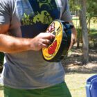 A person in a gray shirt is holding a round BBQ device with a yellow label outdoors. Trees and grass are in the background, indicating a park setting. The person is adjusting or preparing the device.